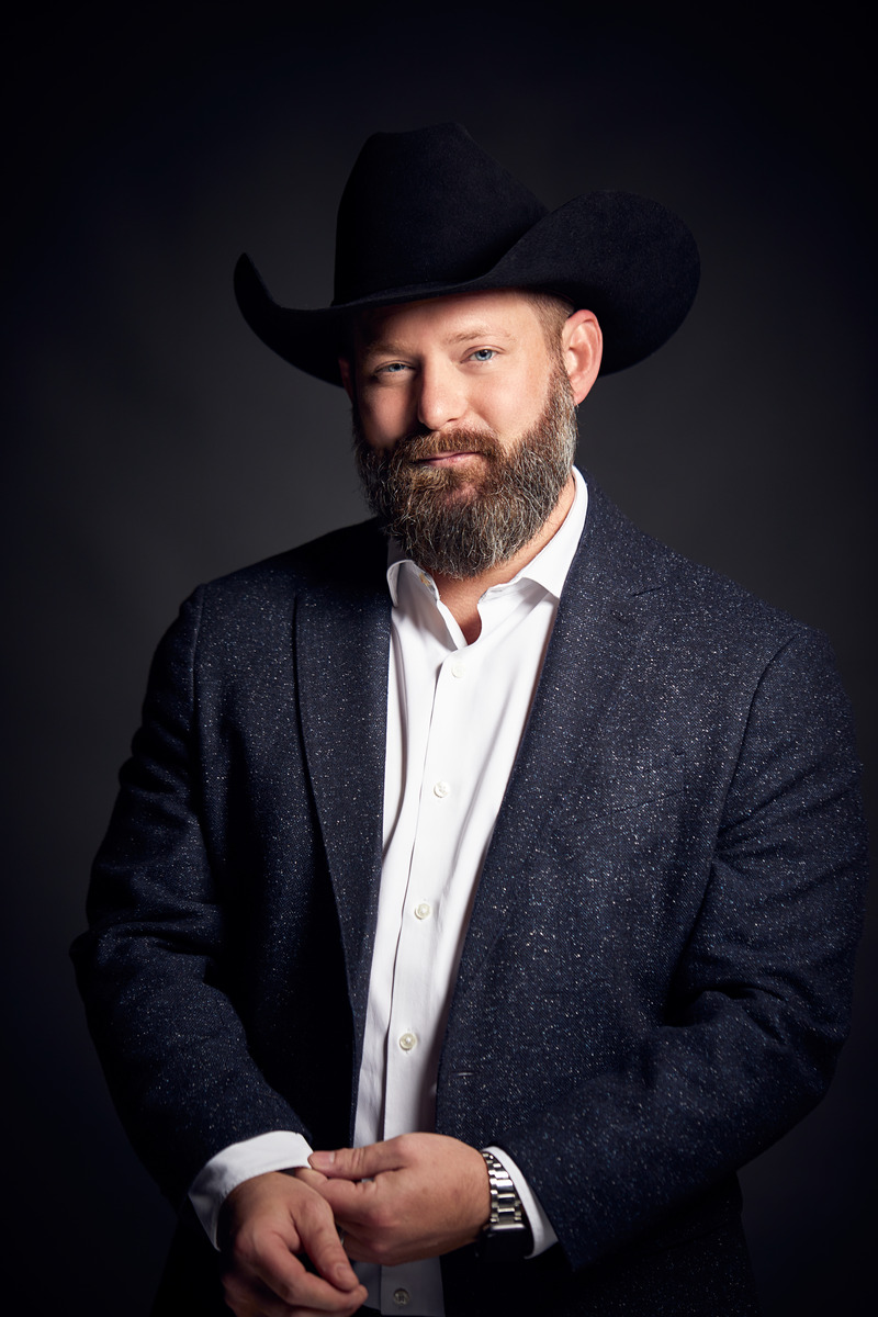 Portrait photo of J.R. Kuhlmann in a dark suit with an open white shirt and black cowboy hat